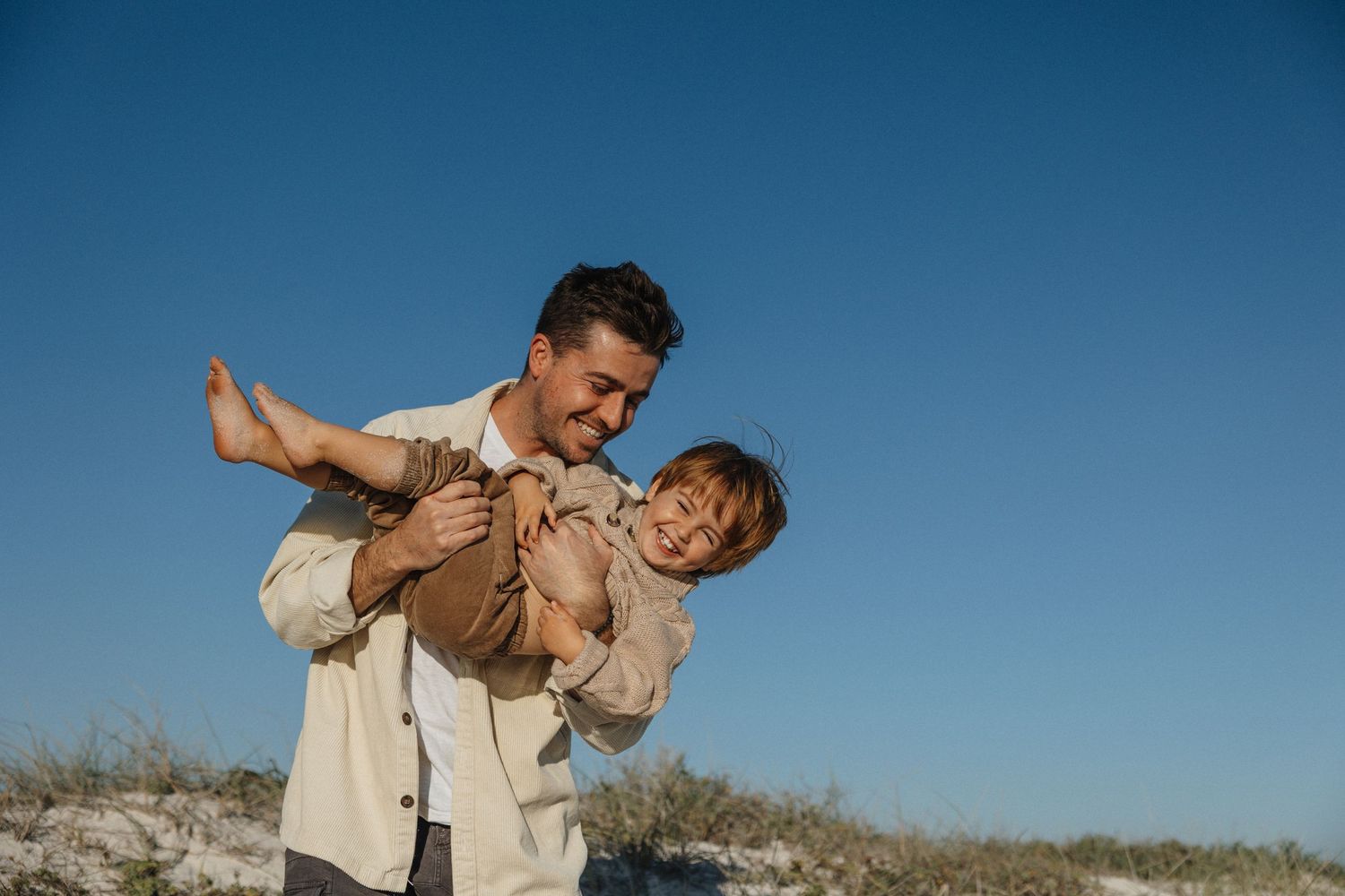 Smiling father playfully lifting laughing child outdoors against bright blue sky on sunny day.