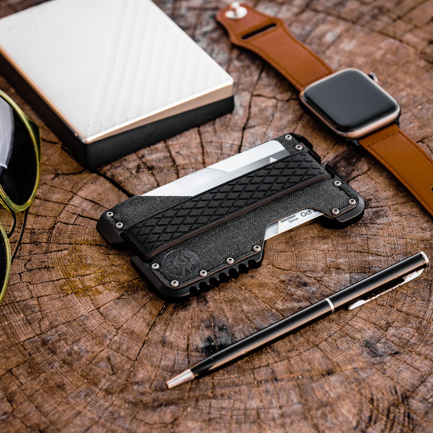 Tactical black metal card wallet on wood surface beside sunglasses, pen, Apple Watch, and external hard drive.