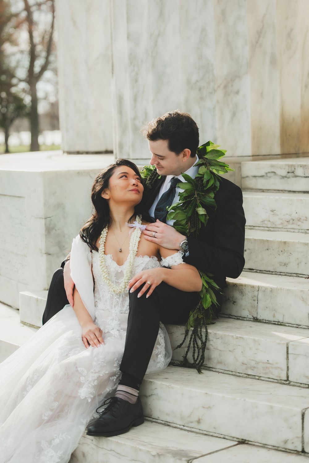 Bride and groom sitting on marble steps, sharing a tender moment, bride in lace gown with pearl necklace, groom in dark suit.