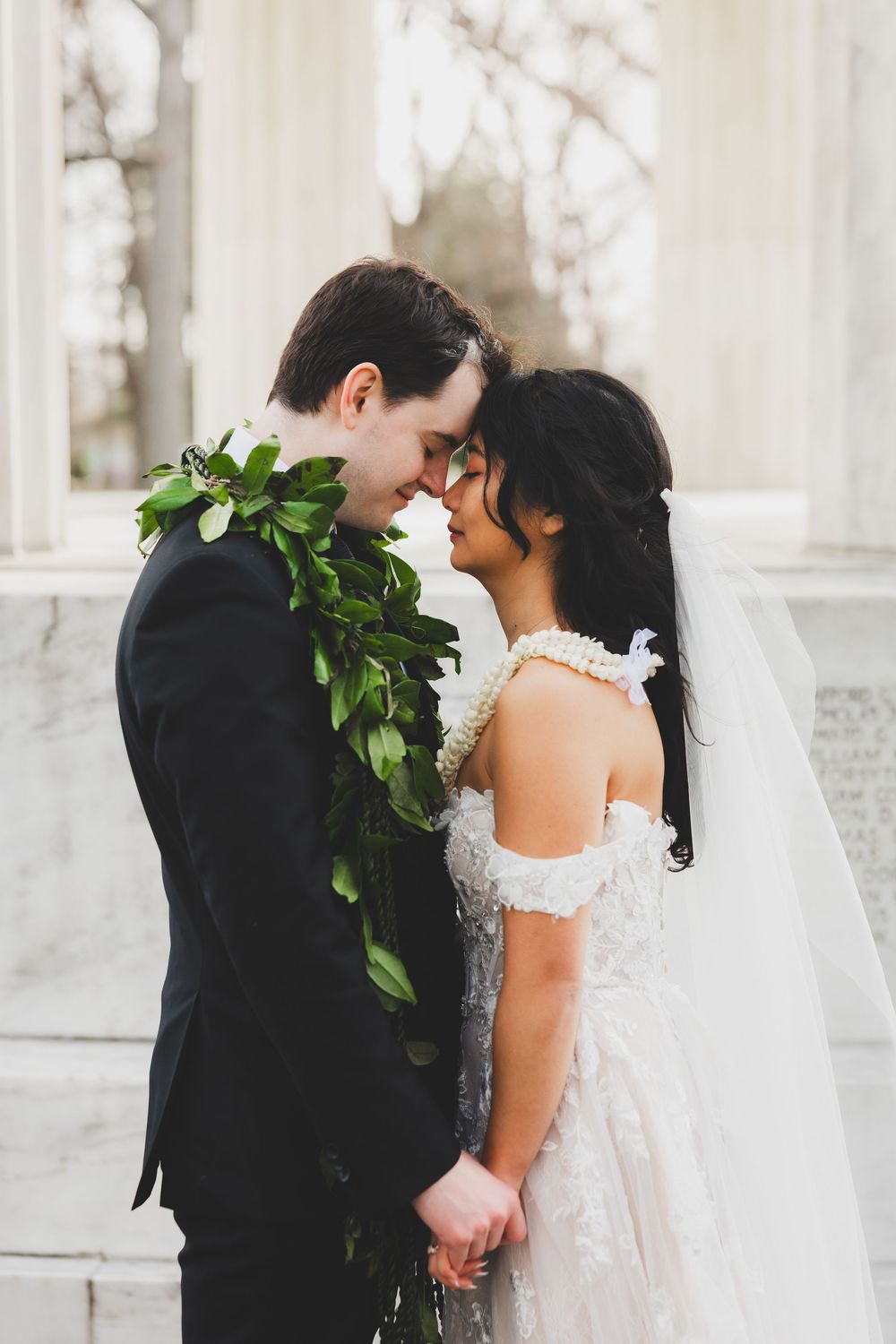 Bride and groom in wedding attire share a romantic moment, wearing floral leis near white marble column for dc wedding portraits