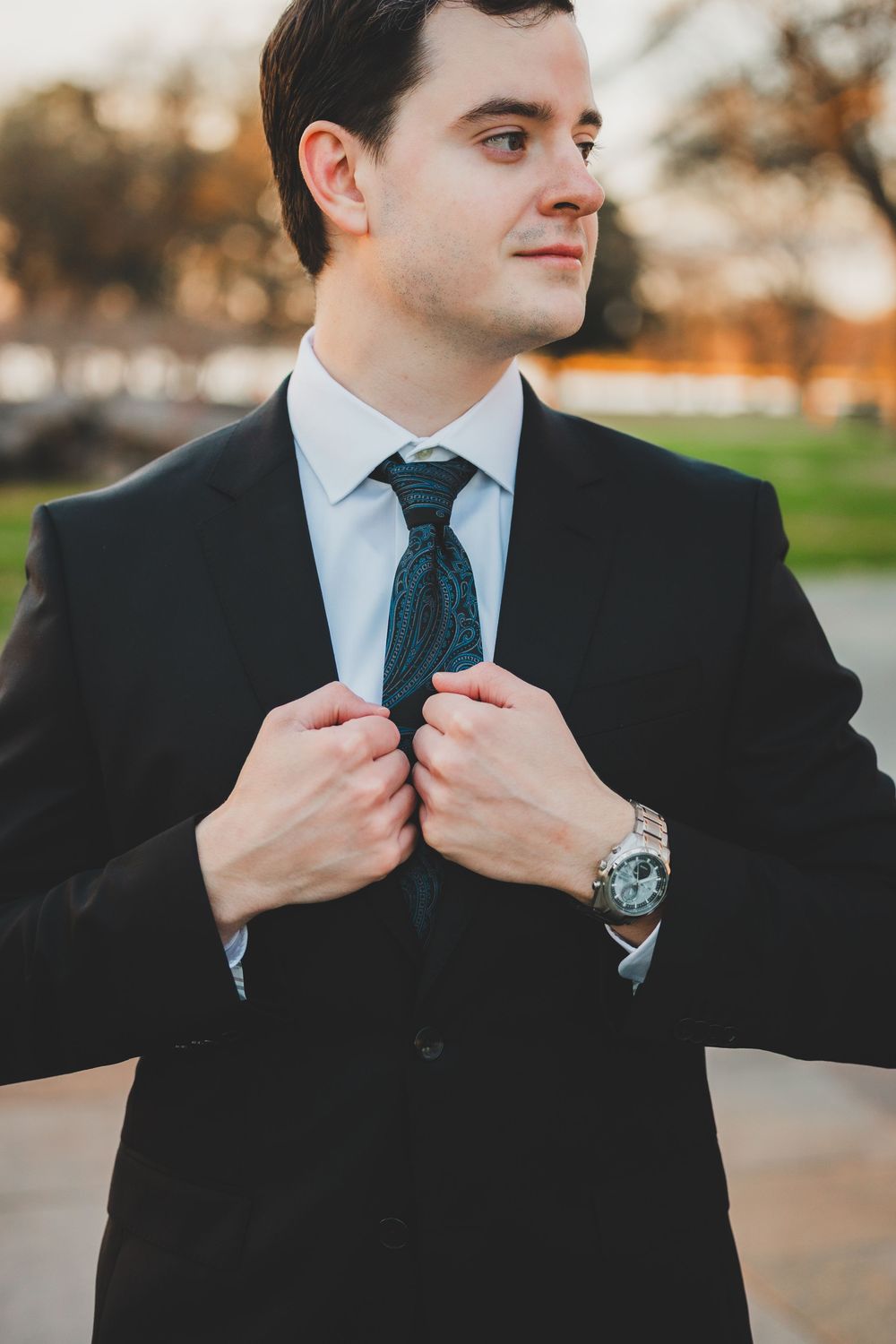 Groom in black suit adjusting tie, wearing silver watch, standing outdoors in warm golden light.