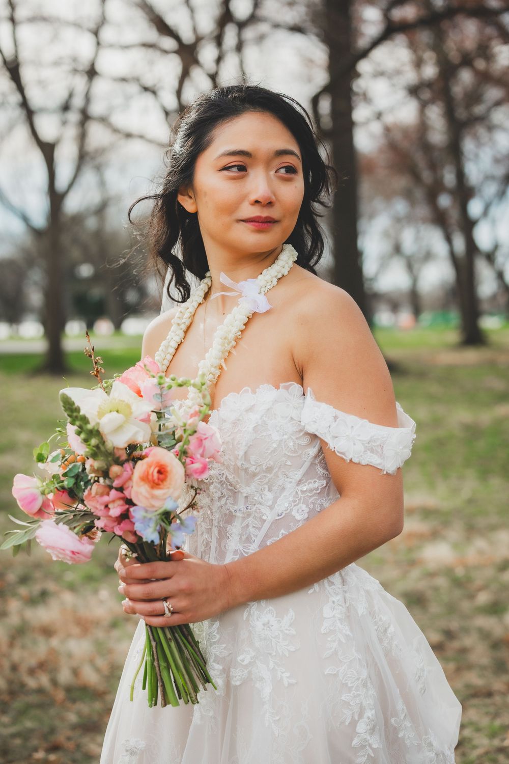 Bride in off-shoulder lace gown holds colorful bouquet of pink roses outdoors among bare trees for a washington dc bridal photo