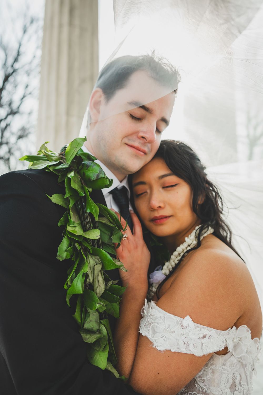 Bride and groom in wedding attire with green lei, sharing a tender embrace with eyes closed under a flowing veil for dc wedding portraits