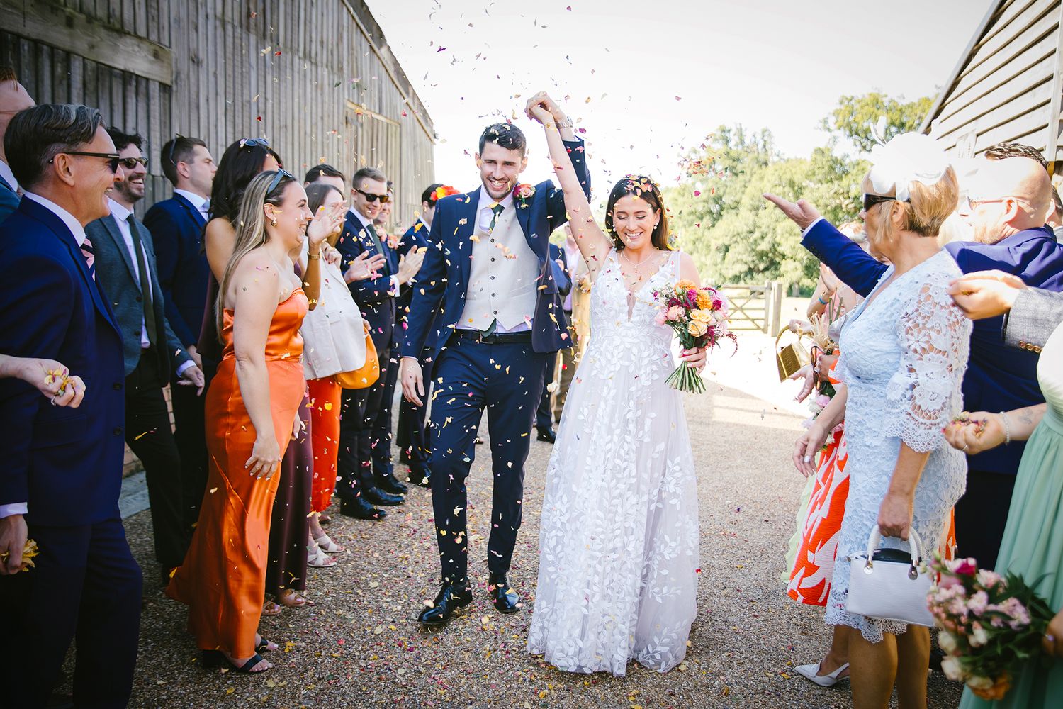 Bride and groom walk through confetti shower surrounded by cheering wedding guests outside a rustic barn venue.