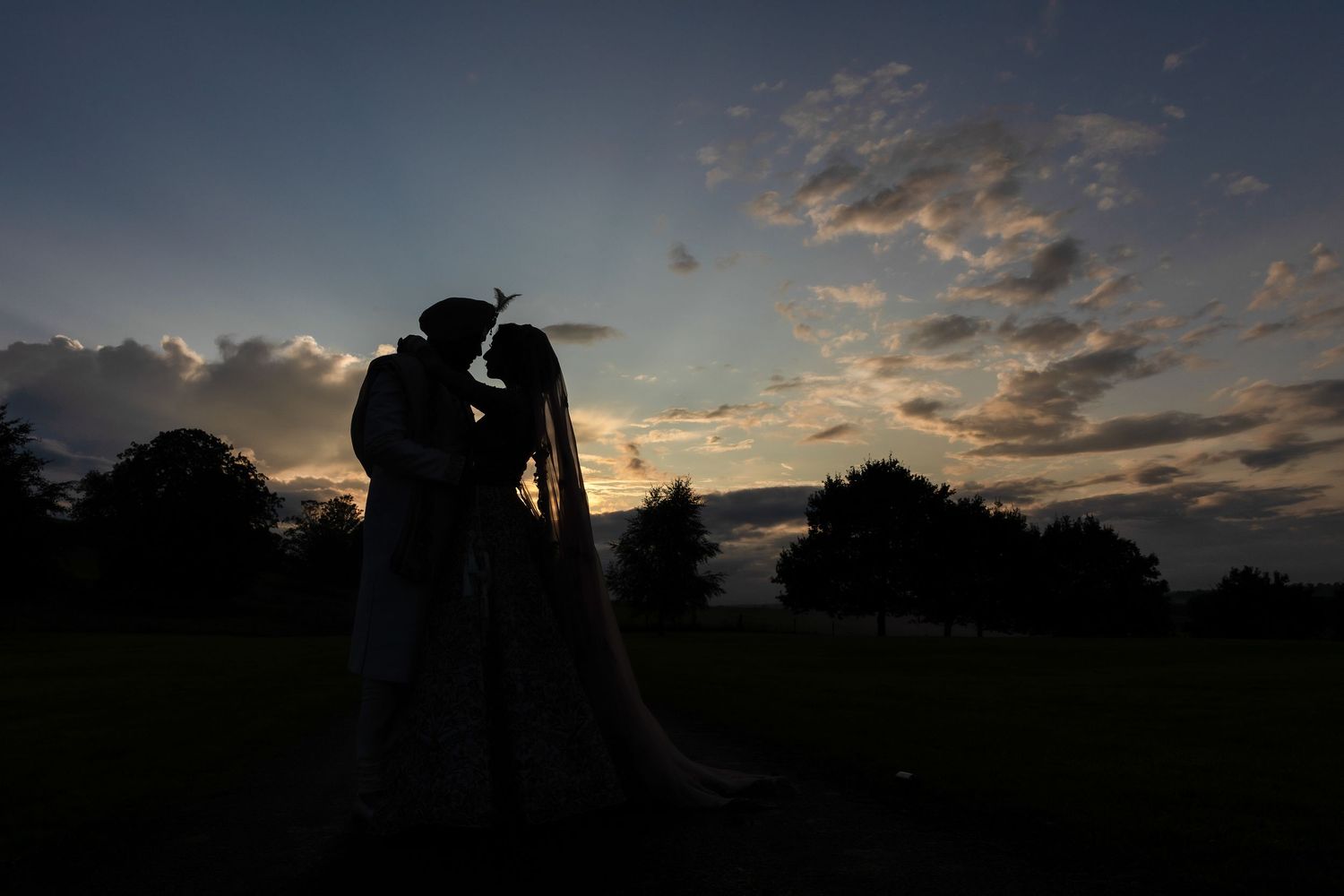 Silhouette of a couple sharing a romantic kiss at sunset with dramatic clouds and trees in the background.
