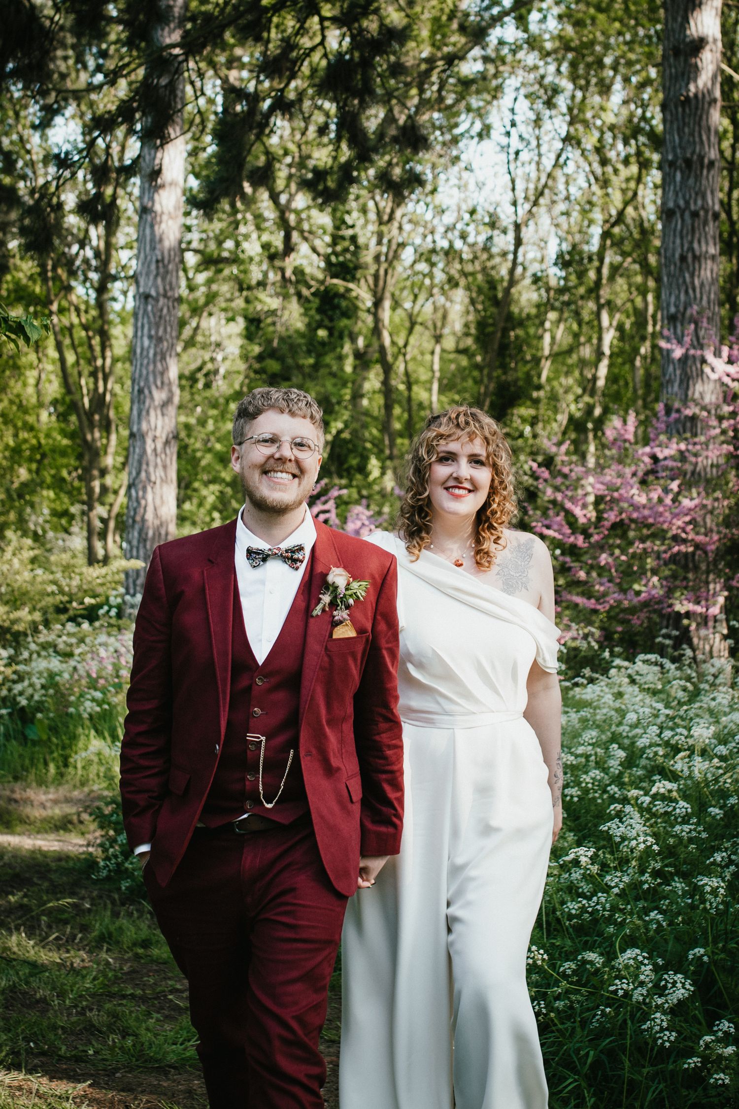 A smiling couple in wedding attire pose in a lush green wooded area at Tipis at Whatton House wedding venue.