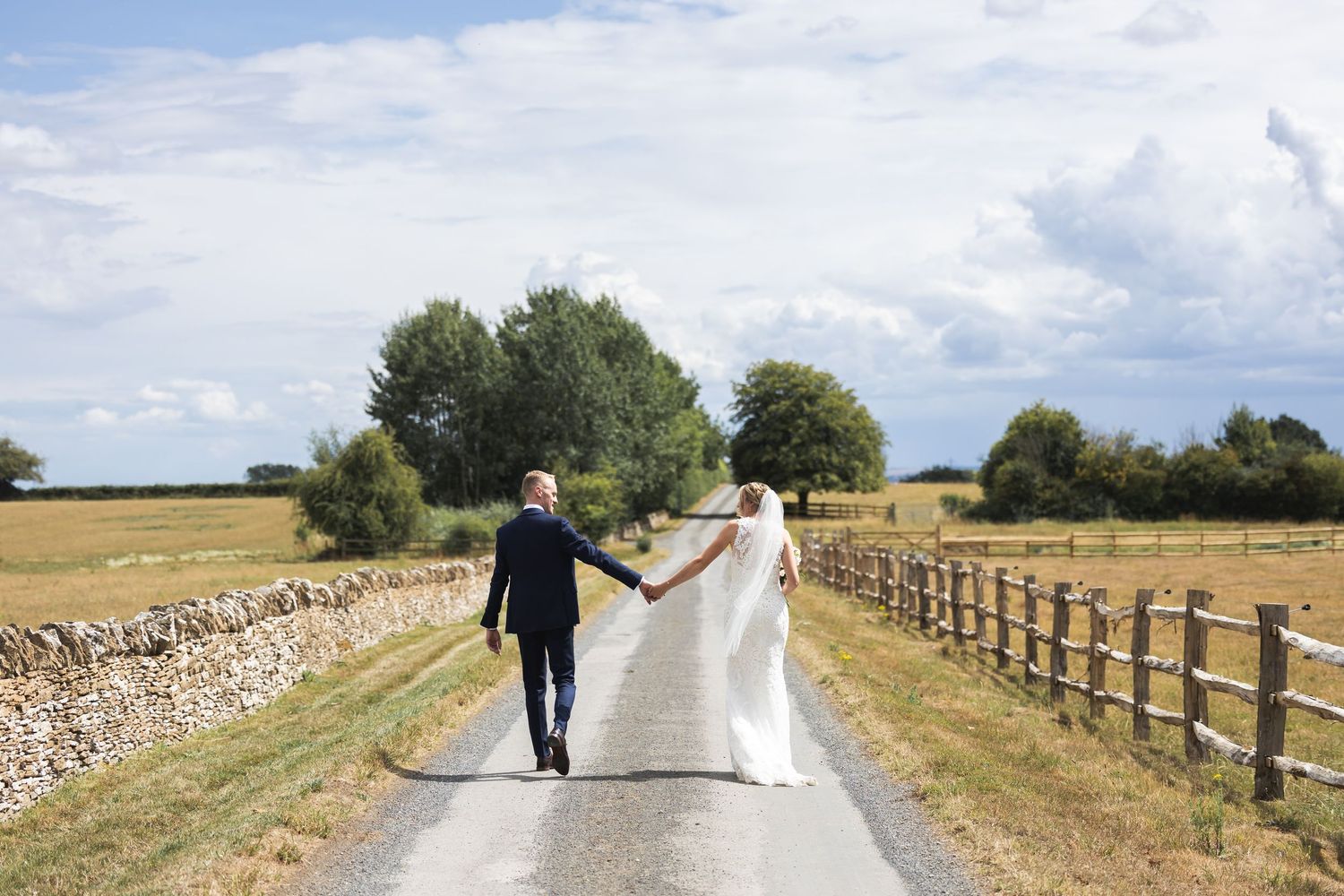 Newlyweds walk hand in hand down Oxleaze Barn's driveway with golden fields and blue sky.