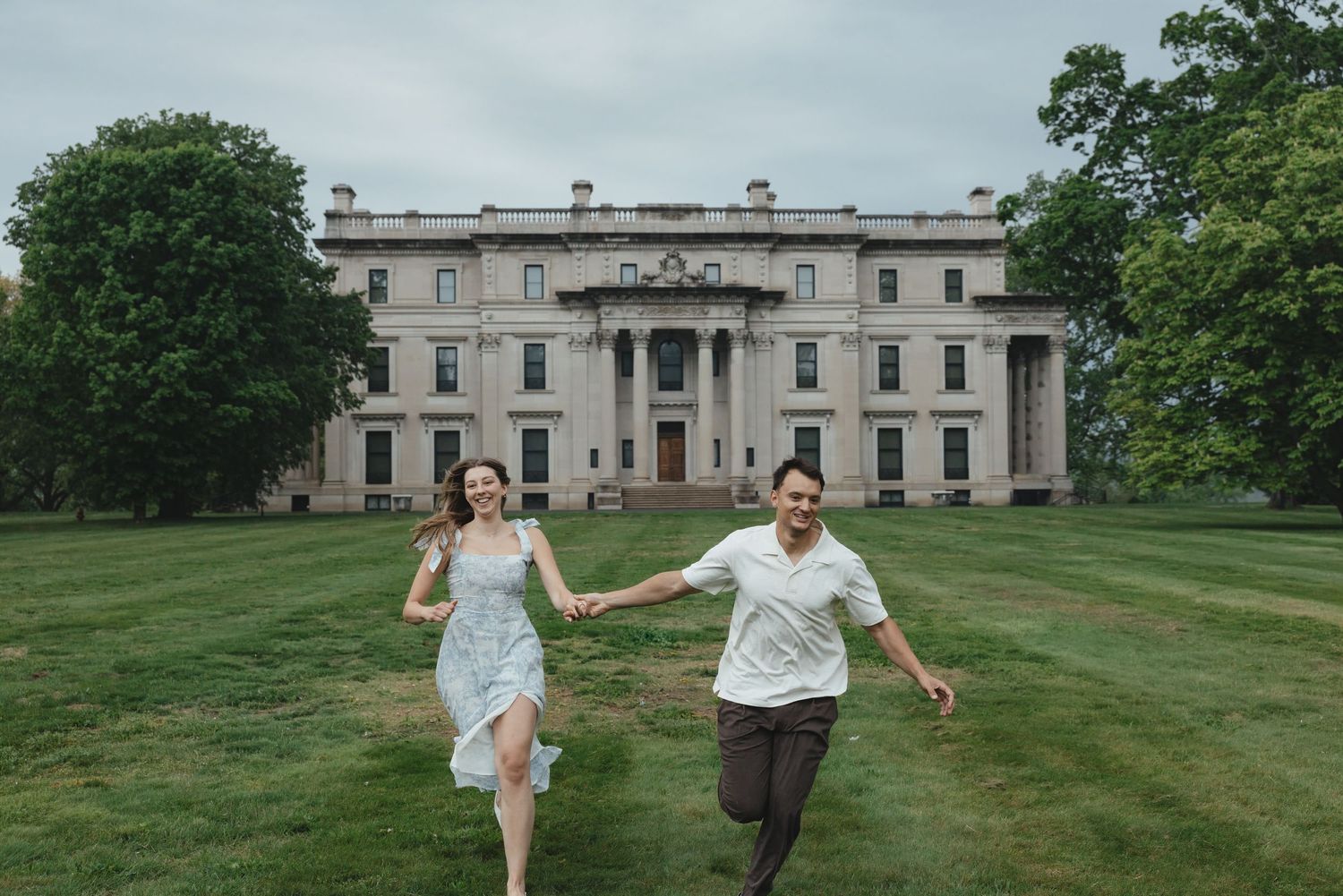A joyful couple runs hand-in-hand across a lush green lawn in front of a grand historic mansion.
