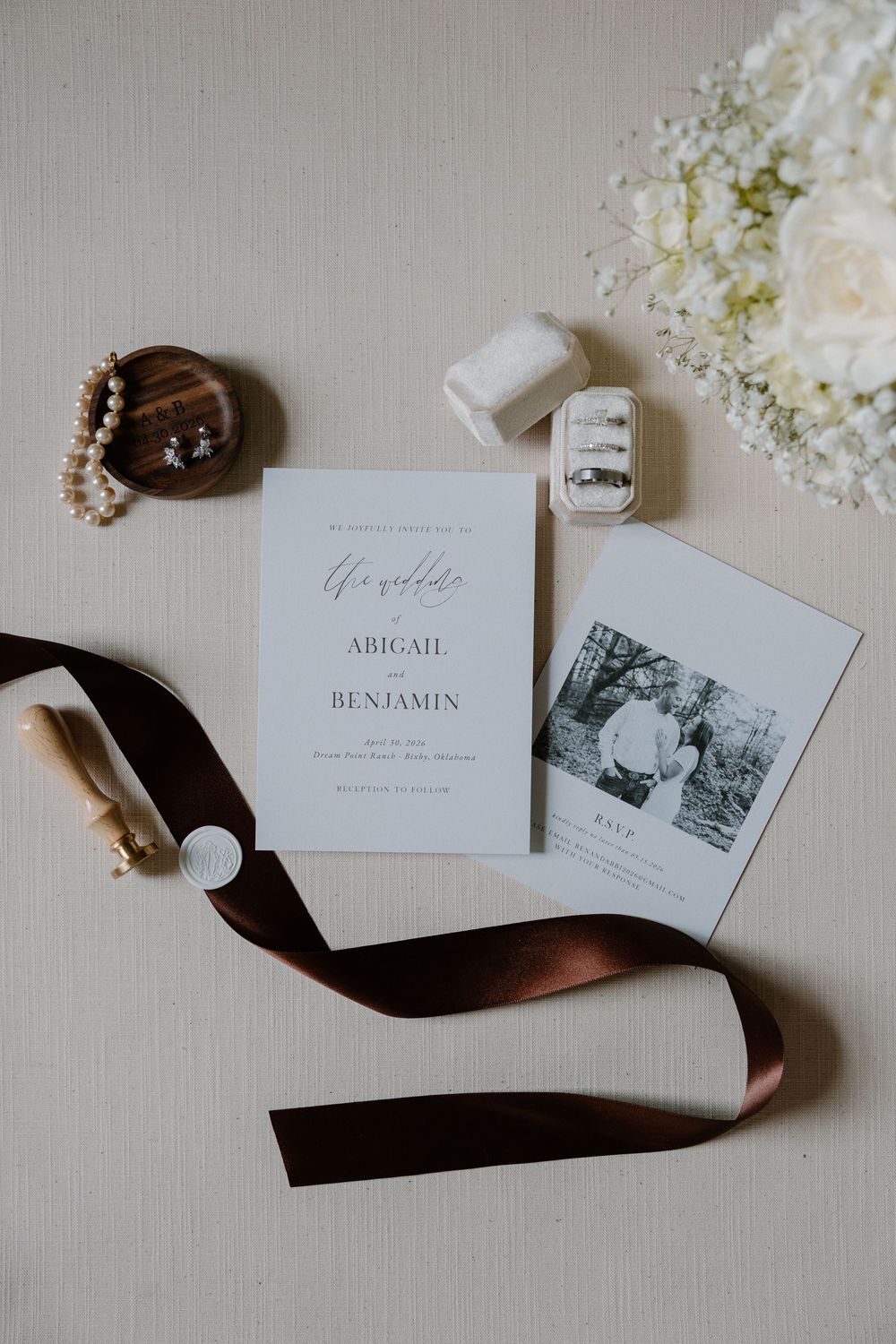 Elegant wedding stationery flatlay with invitation, rings, pearl jewelry, and white floral bouquet.