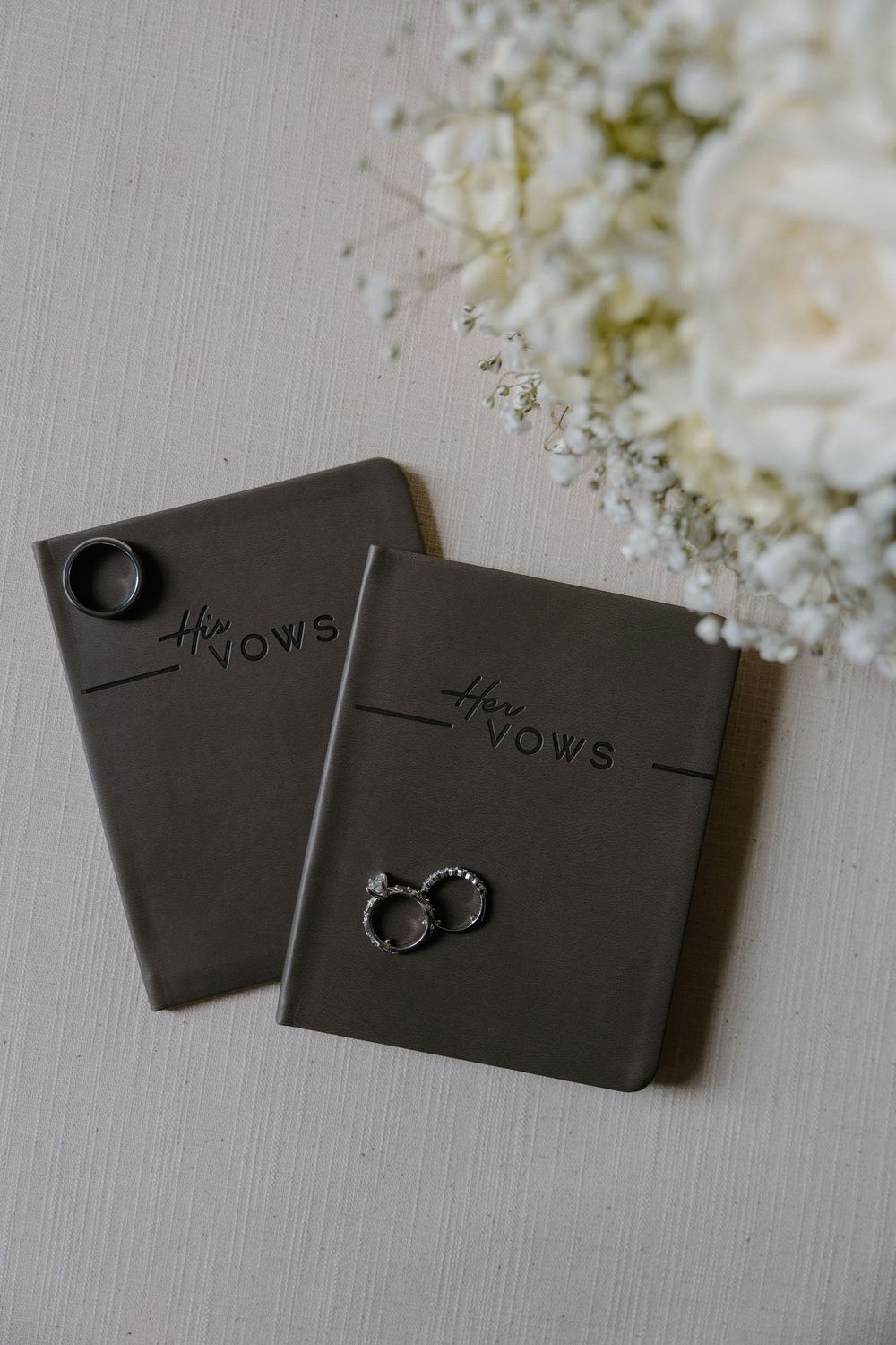 His and hers dark leather vow books with wedding rings resting on them beside a white floral bouquet.