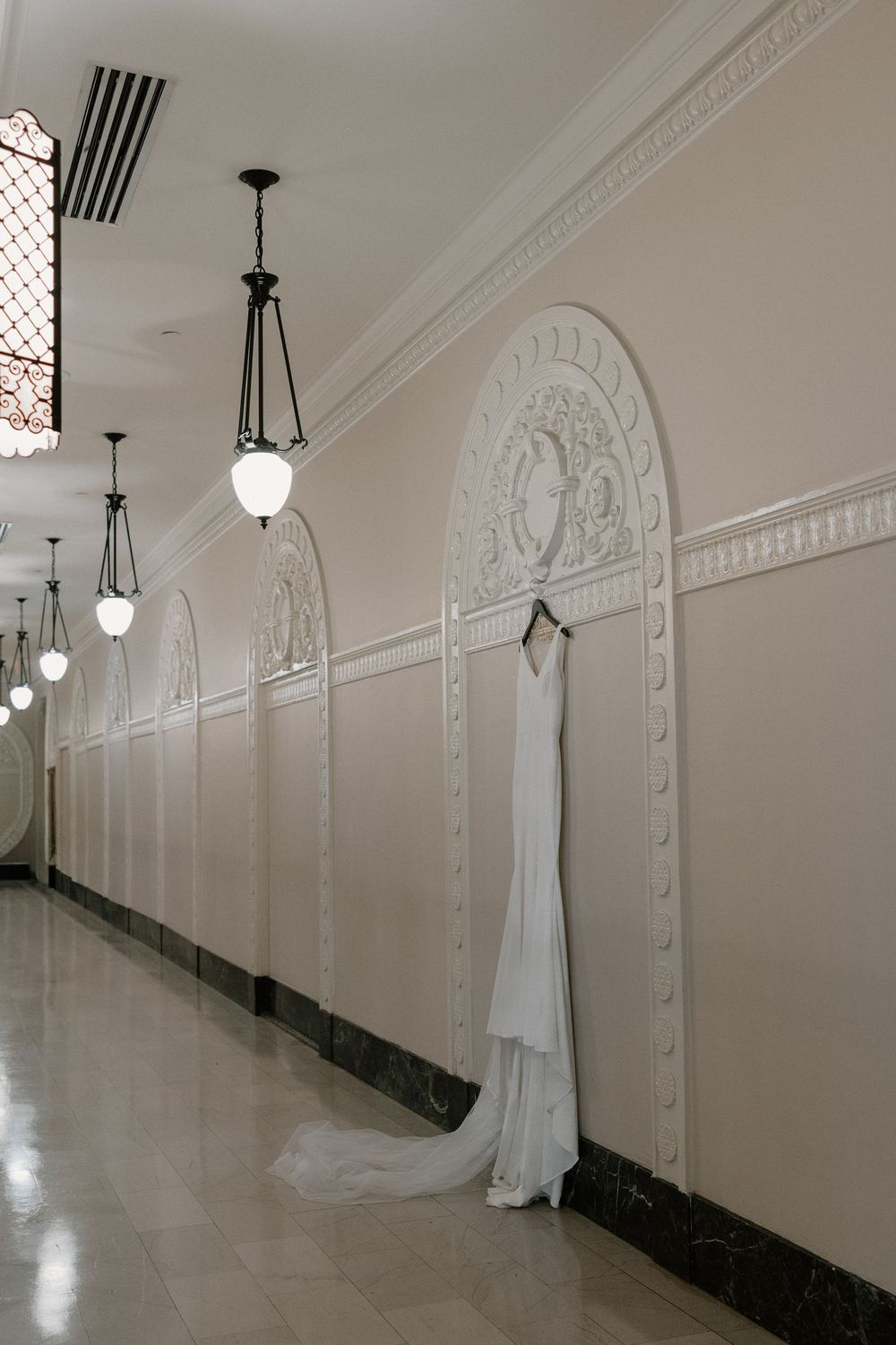 White wedding gown hanging in an elegant hallway with ornate arched walls and vintage pendant lights.