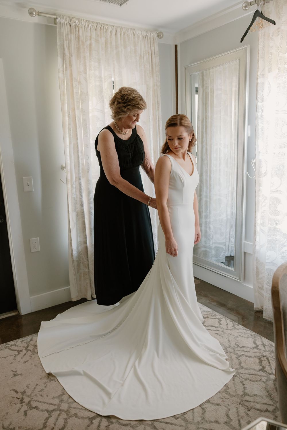 Mother helping bride button her elegant white wedding gown with long train before the ceremony.