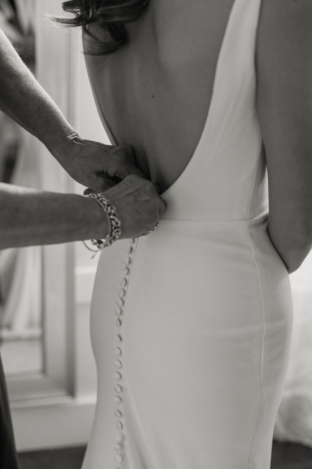 Black and white close-up of hands fastening delicate buttons on backless bridal wedding gown.