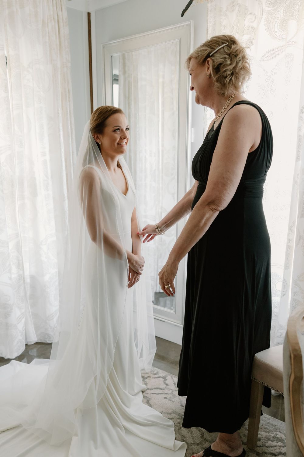 Bride in white gown and veil holds hands with woman in black dress during wedding preparations.