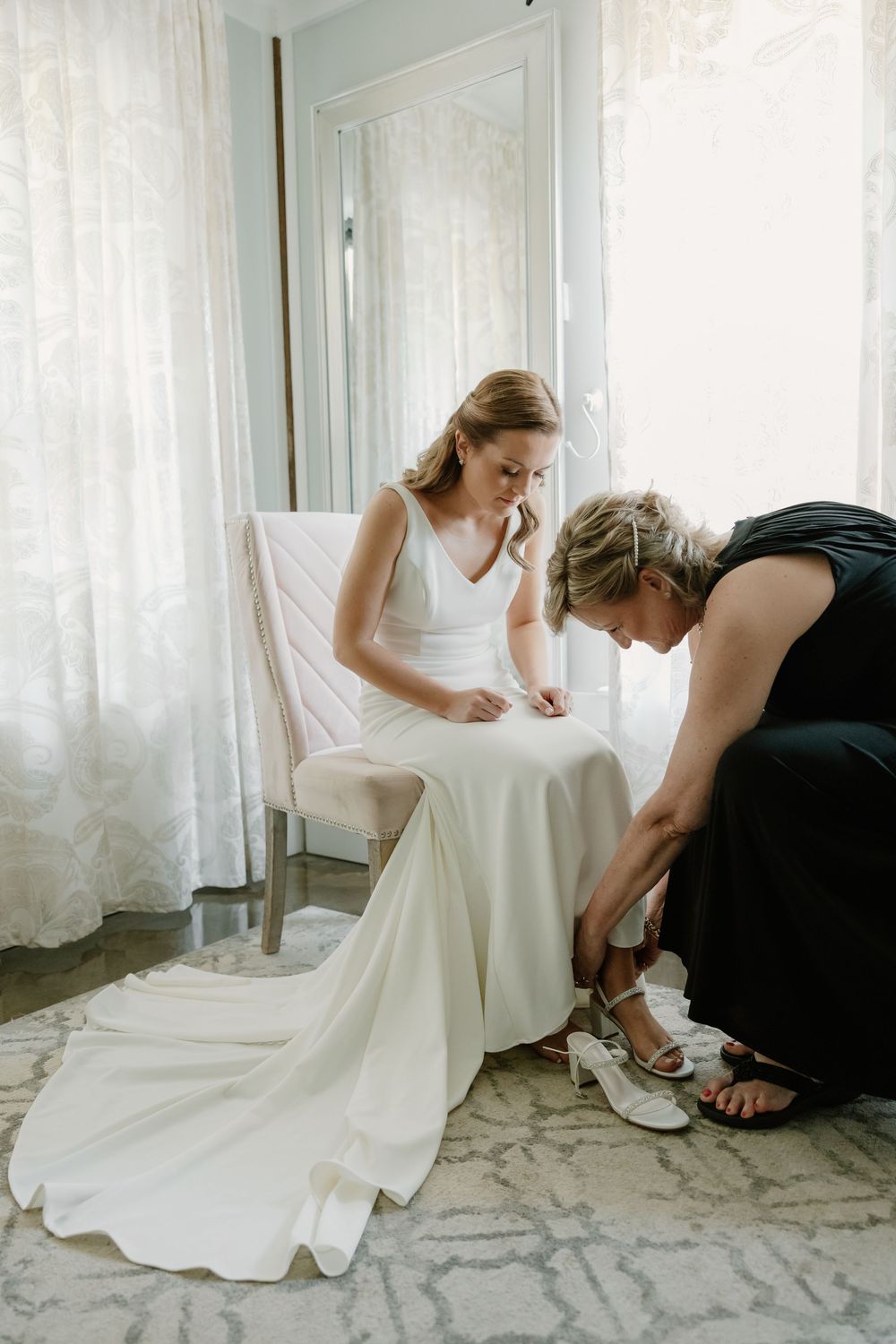A bride sits as her mother helps put on her wedding shoes in a bright, airy bridal suite.