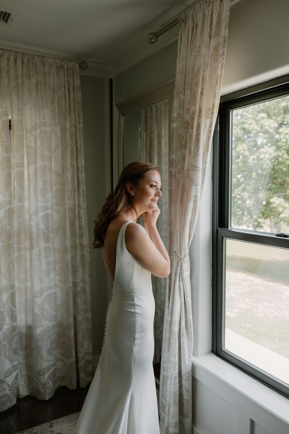 Bride in elegant white gown gazing out window with sheer curtains on her wedding day.
