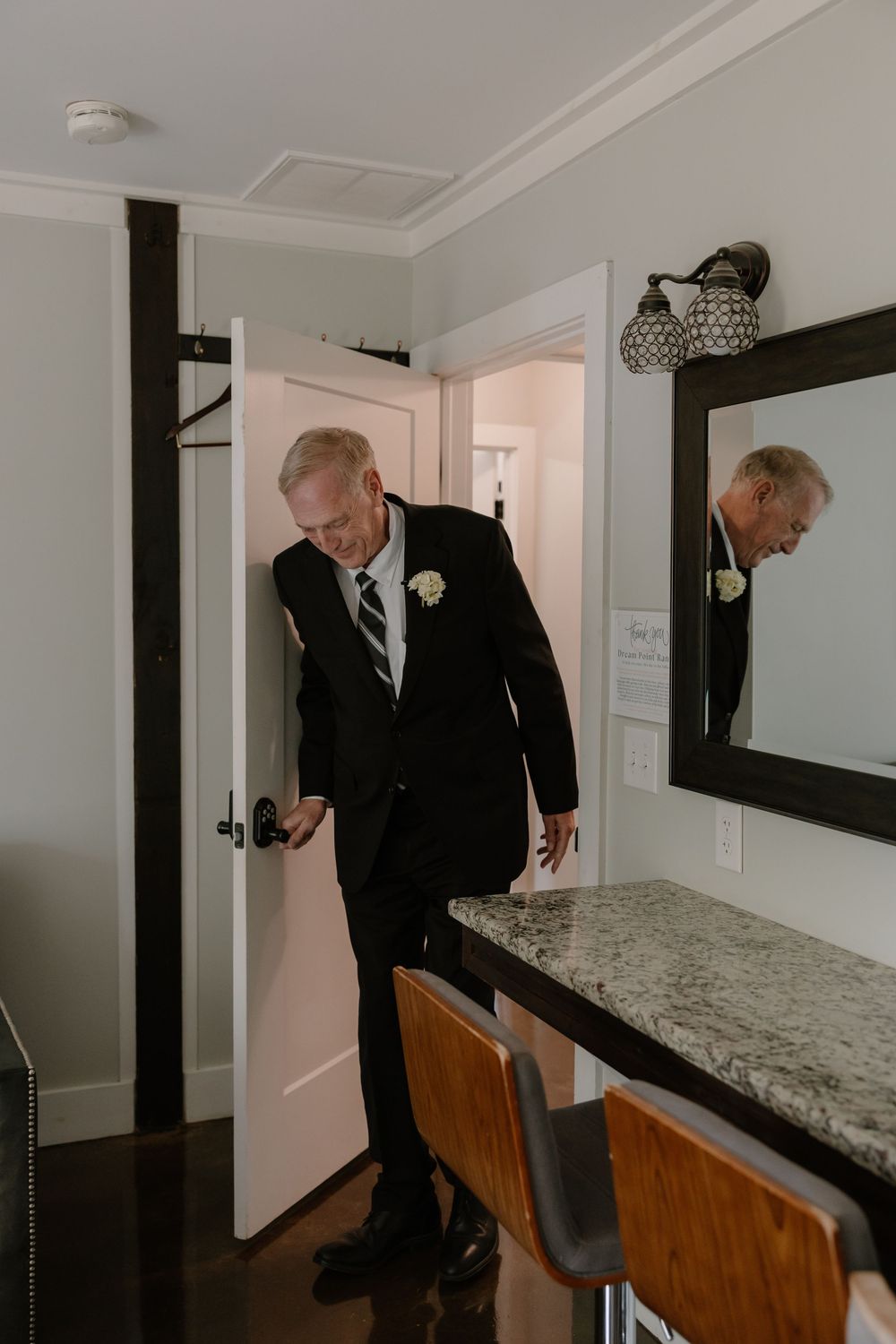 Elderly man in black suit with boutonniere entering hotel room door, reflected in nearby wall mirror.