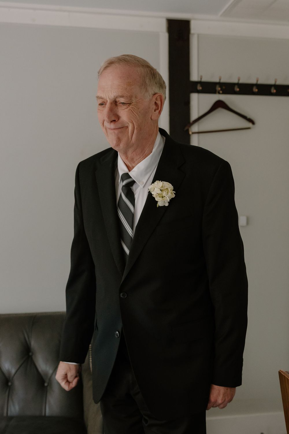 Elderly man in black suit with white boutonniere and striped tie smiling at a wedding.