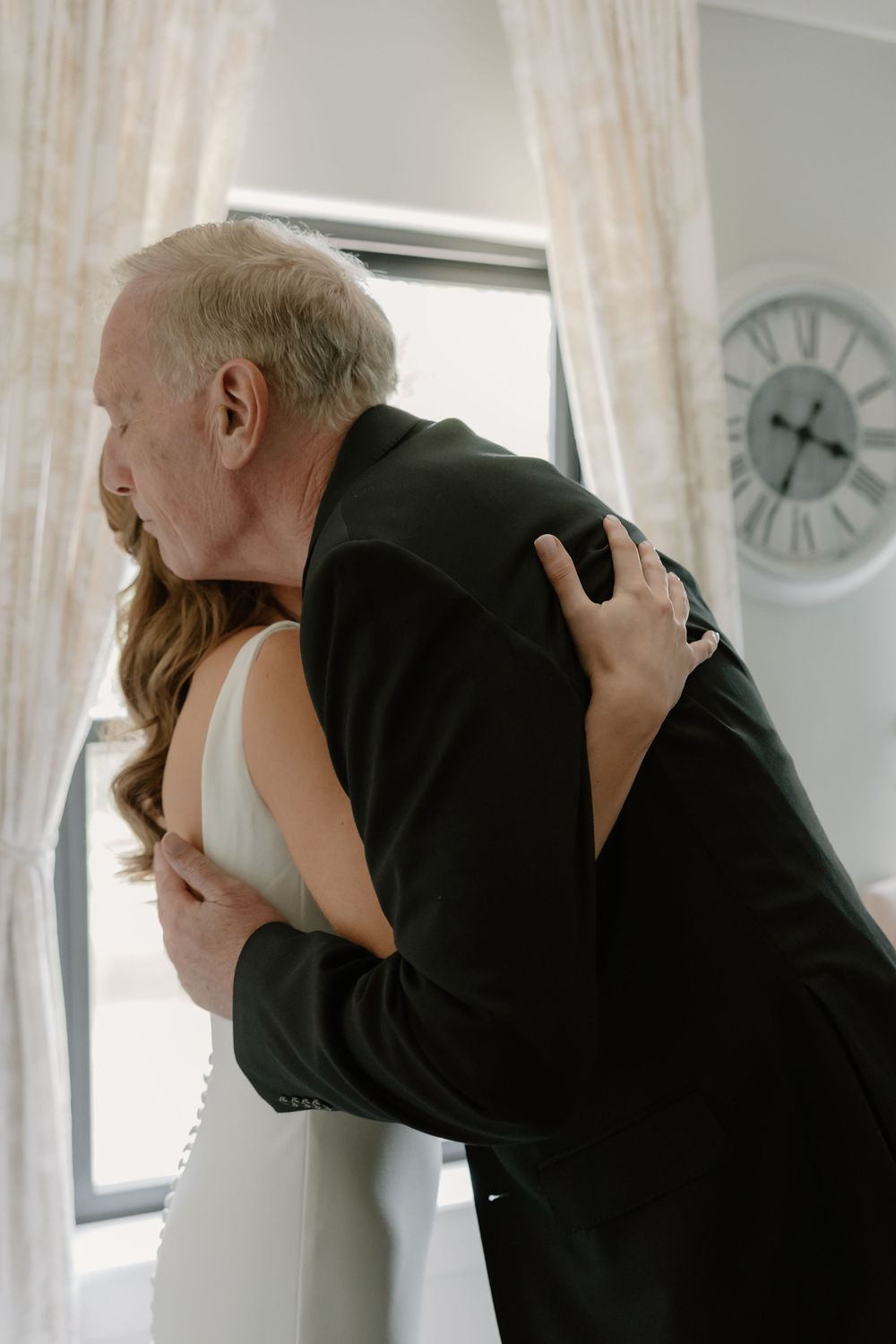 Father and bride sharing an emotional hug in a bright room with a clock on the wall on wedding day.
