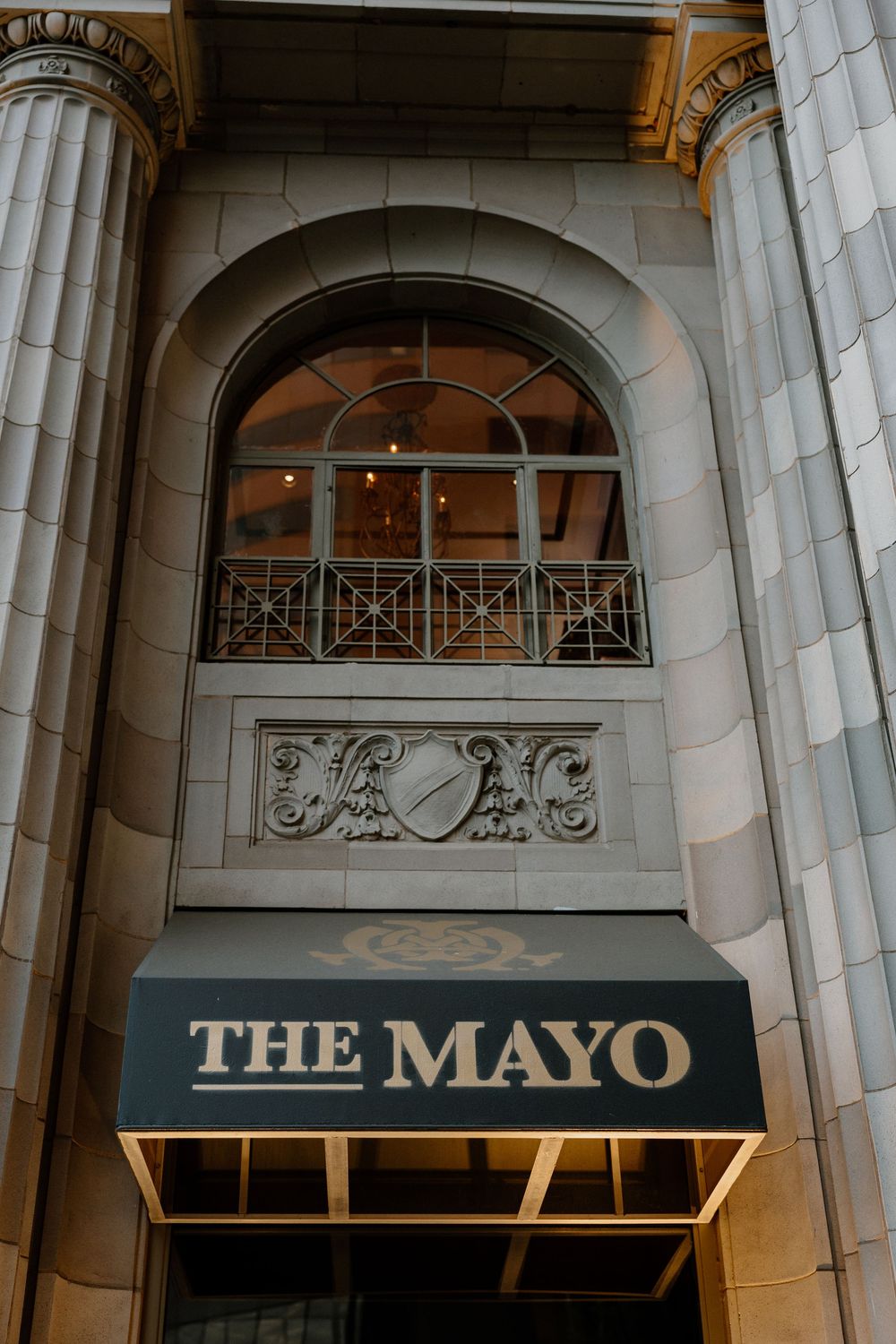 The Mayo Hotel entrance featuring elegant stone architecture, arched window, ornate crest detail, and gold-trimmed black awning.