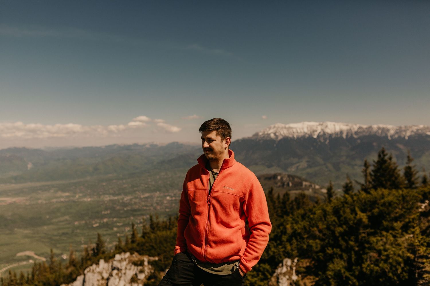 Man in red jacket standing on mountain summit with snow-capped peaks and valley views in background.