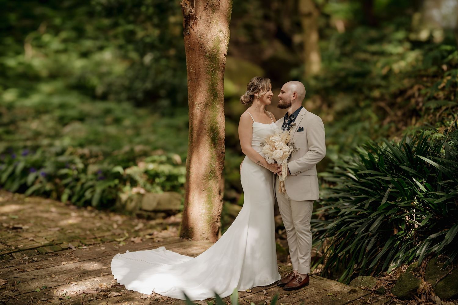Bride and groom share a romantic moment in a lush green forest setting, bride in a fitted white gown holding a bouquet.