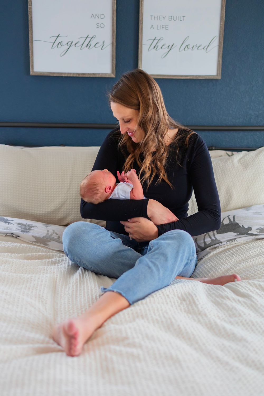A smiling mother sits cross-legged on a bed cradling her newborn baby, with motivational wall art behind her.