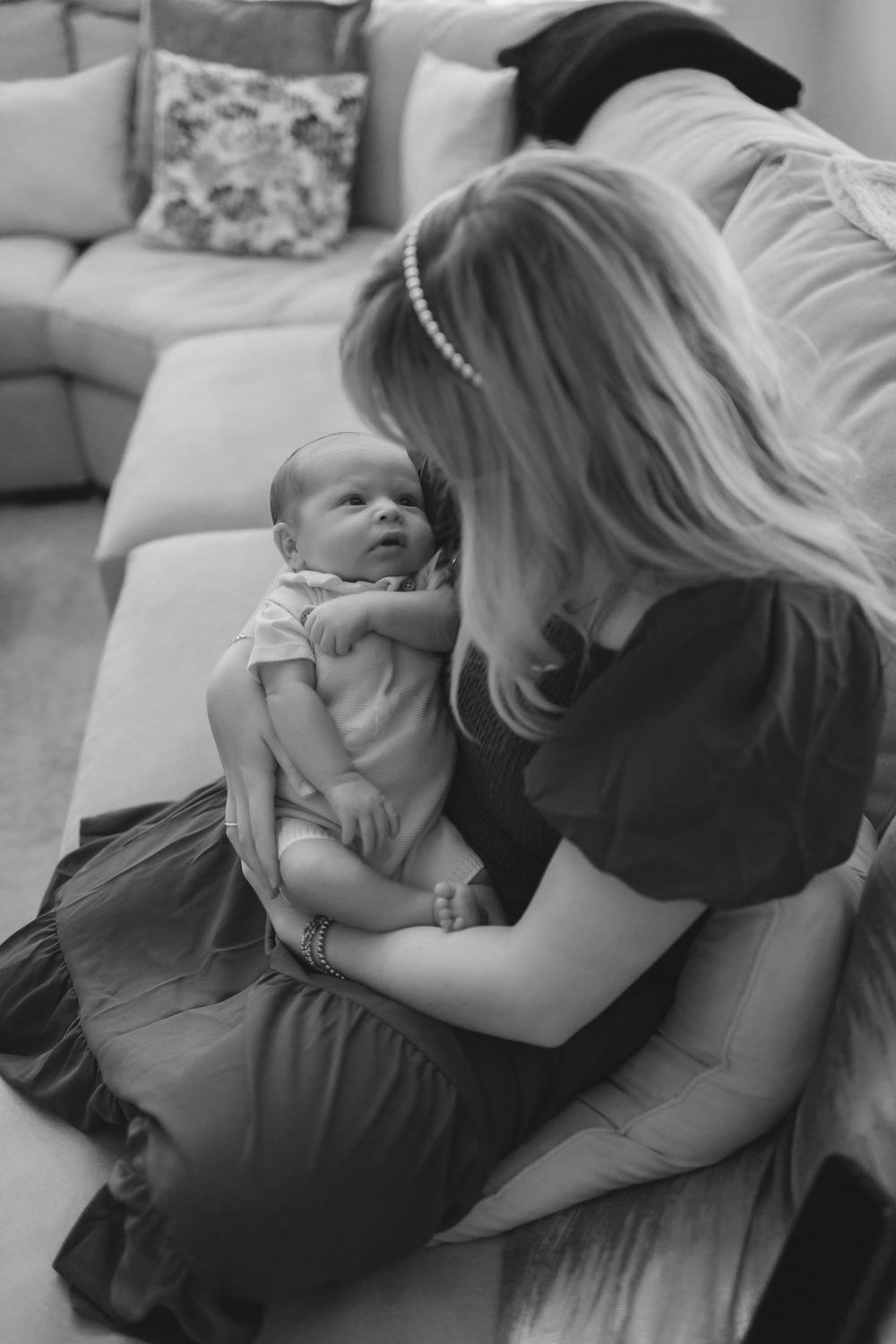 Black and white photo of a mother sitting on sofa holding her newborn baby, gazing lovingly at each other indoors.