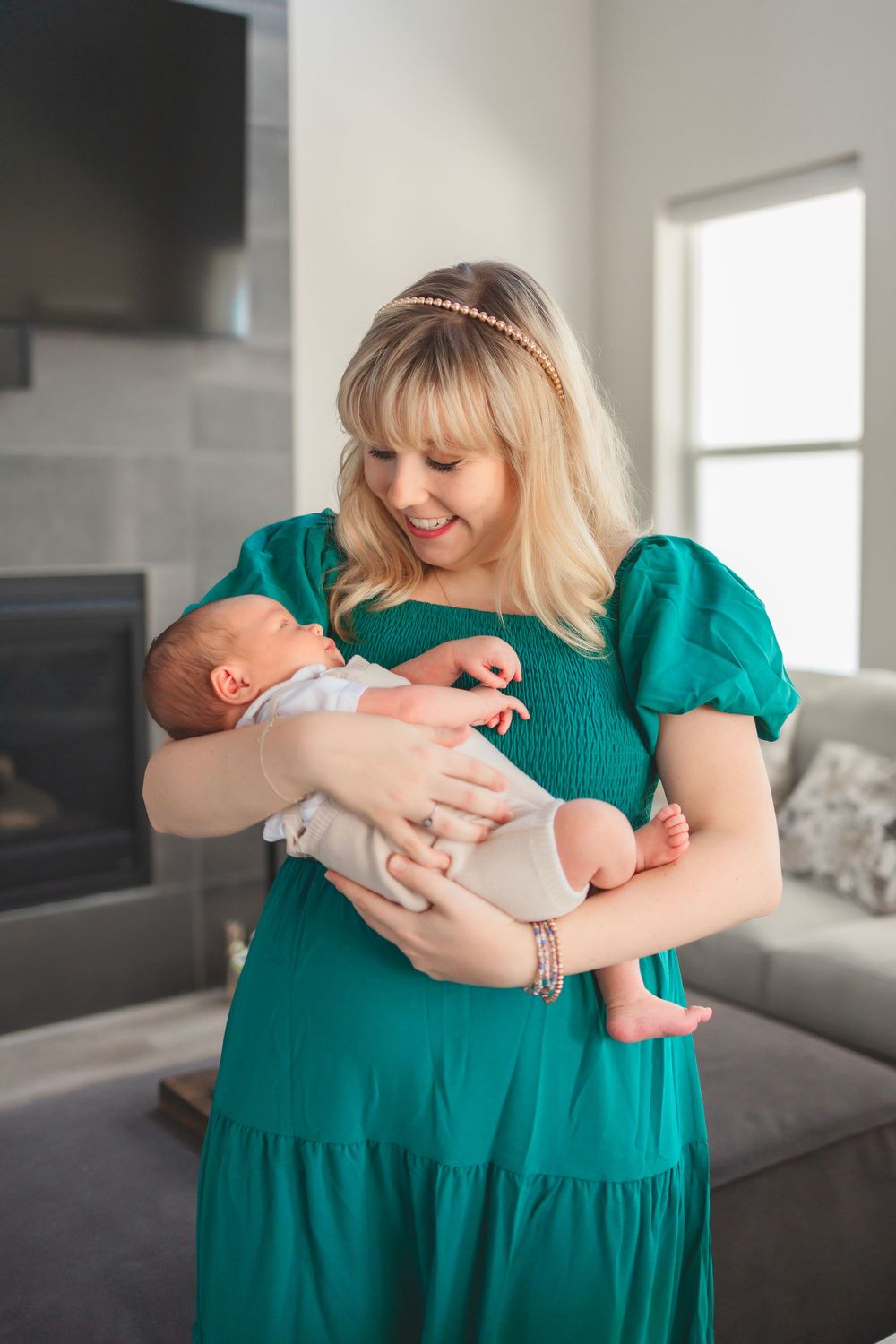 Smiling blonde woman in teal dress lovingly holding her newborn baby in a modern living room.
