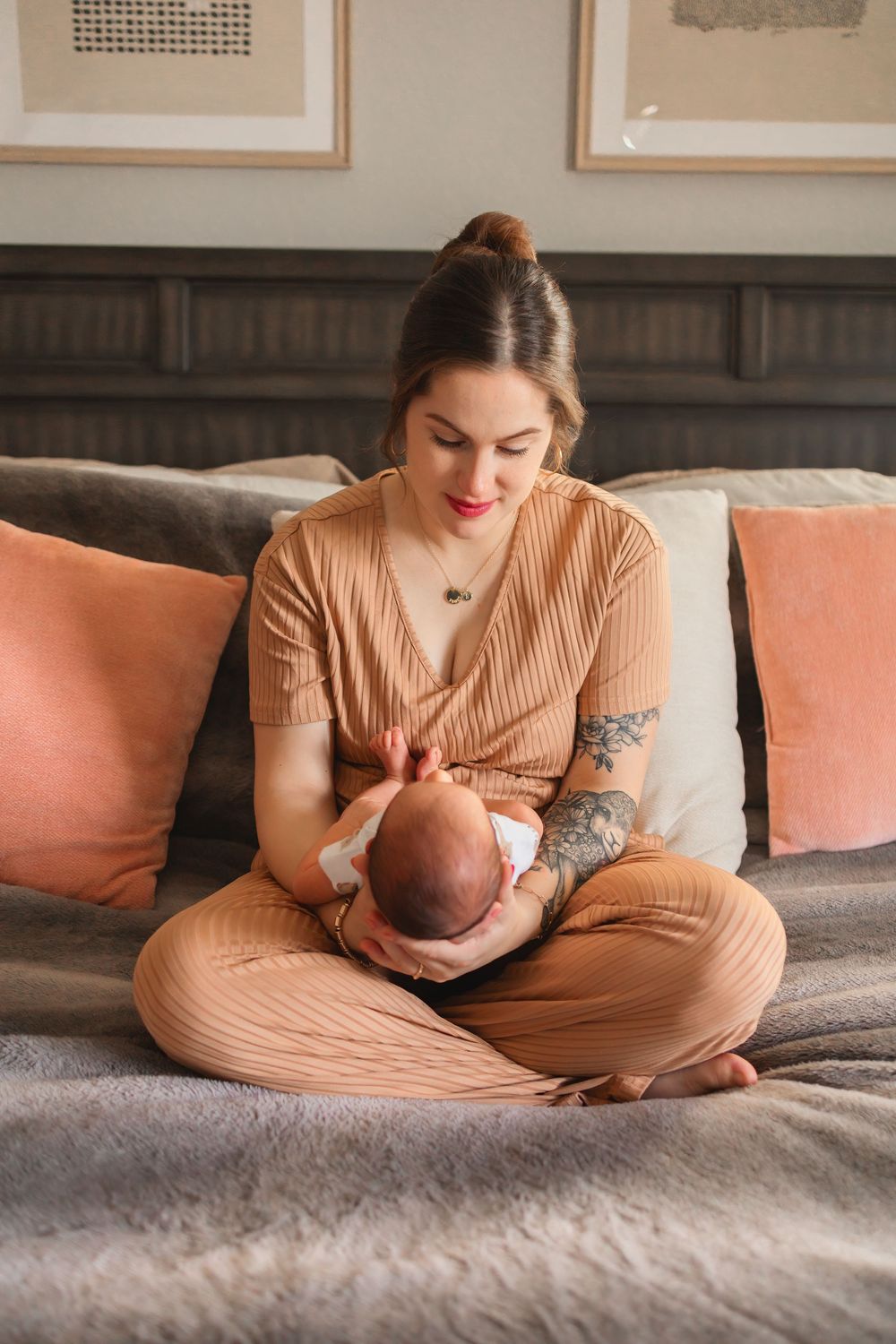 Mother sits cross-legged on bed cradling newborn baby, wearing tan outfit with pink pillows behind her.