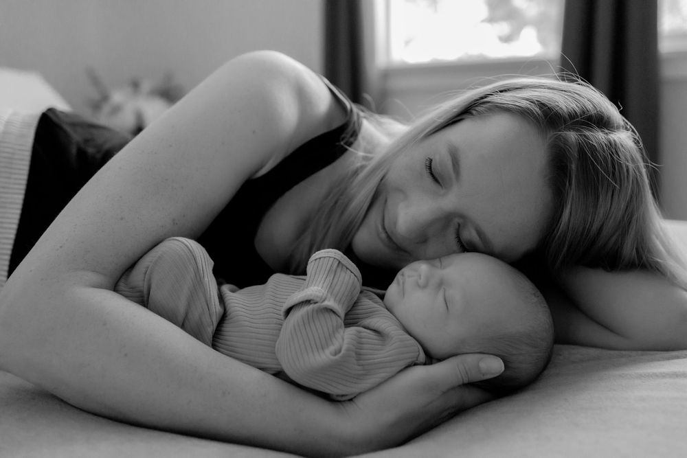 Black and white photo of a mother tenderly cuddling her sleeping newborn baby on a bed.
