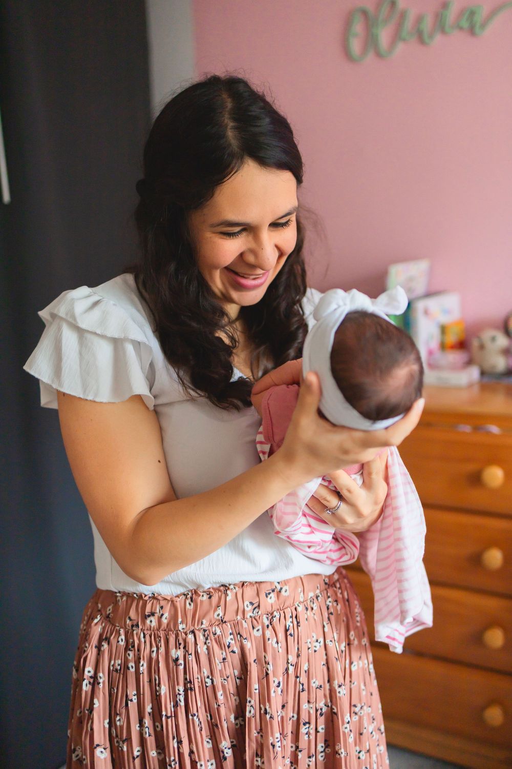 Smiling mother holding newborn baby in pink nursery room with Olivia name sign on wall.