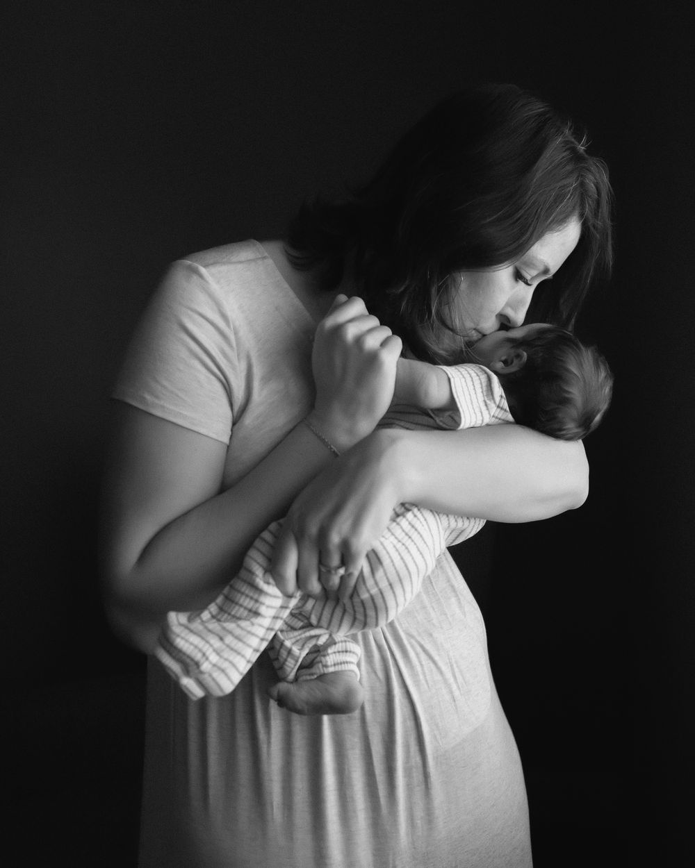 Black and white photo of a mother tenderly kissing her newborn baby while cradling them in her arms.