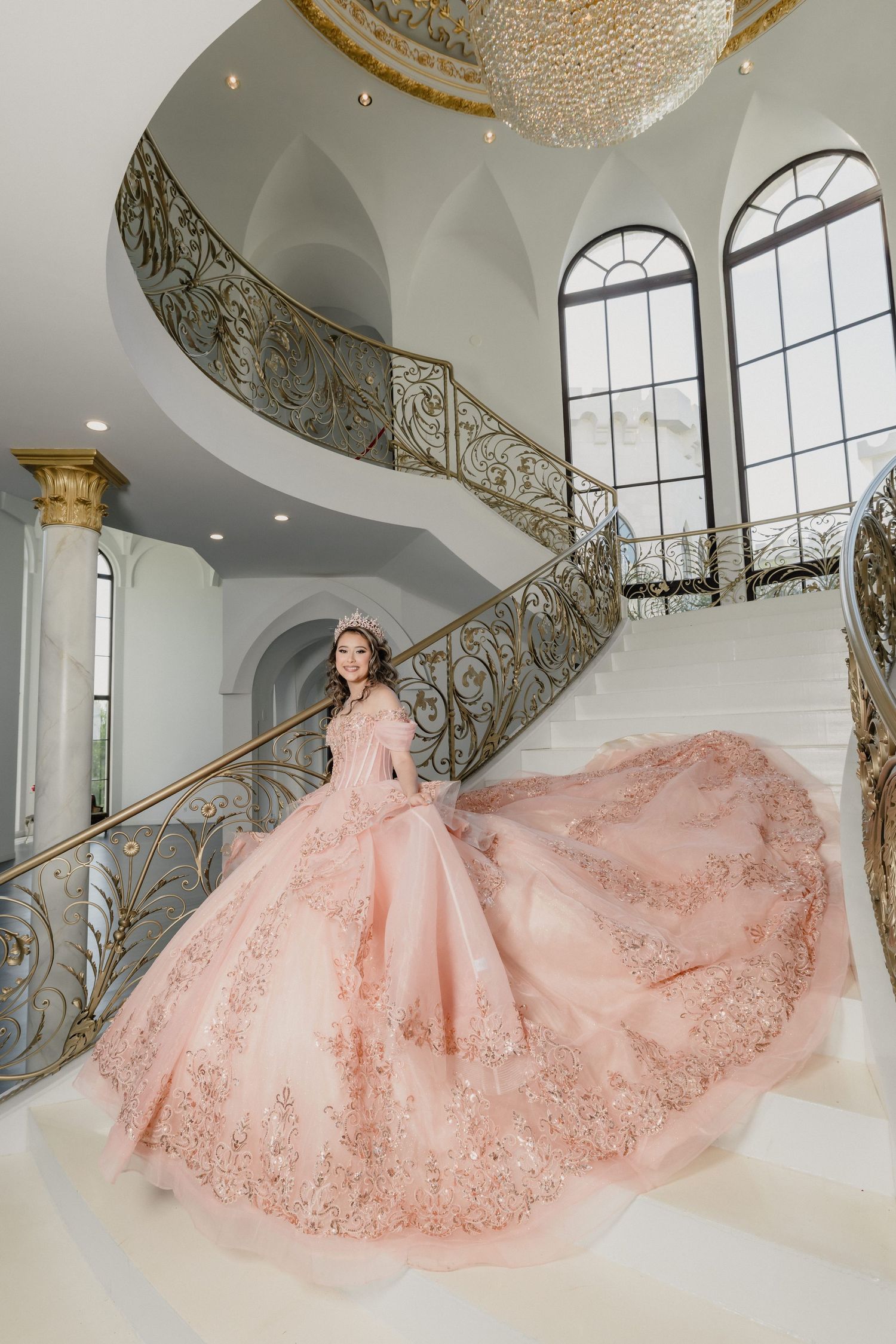 Young woman in pink quinceañera ball gown with lace details poses on grand spiral staircase in elegant mansion.