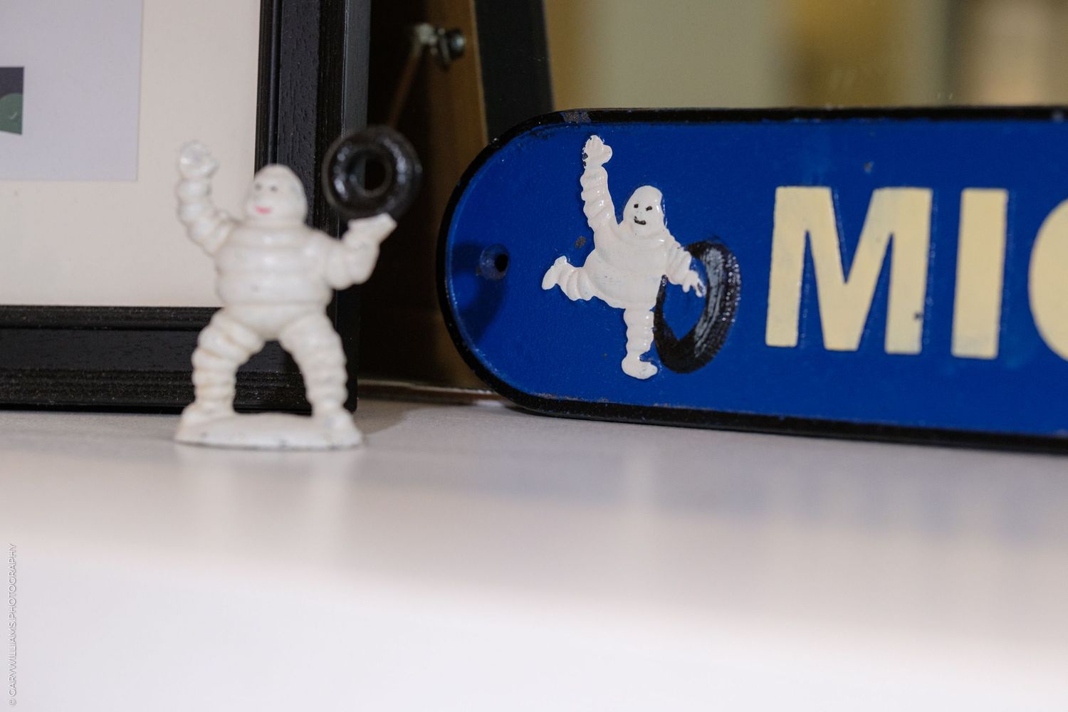 White Michelin Man figurine beside a vintage Michelin tire brand sign on a white shelf.