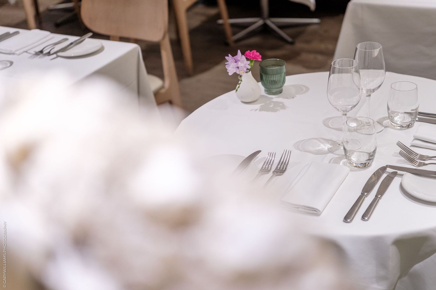 Fine dining table set with wine glasses, silverware, white linens, and small pink flower vase.