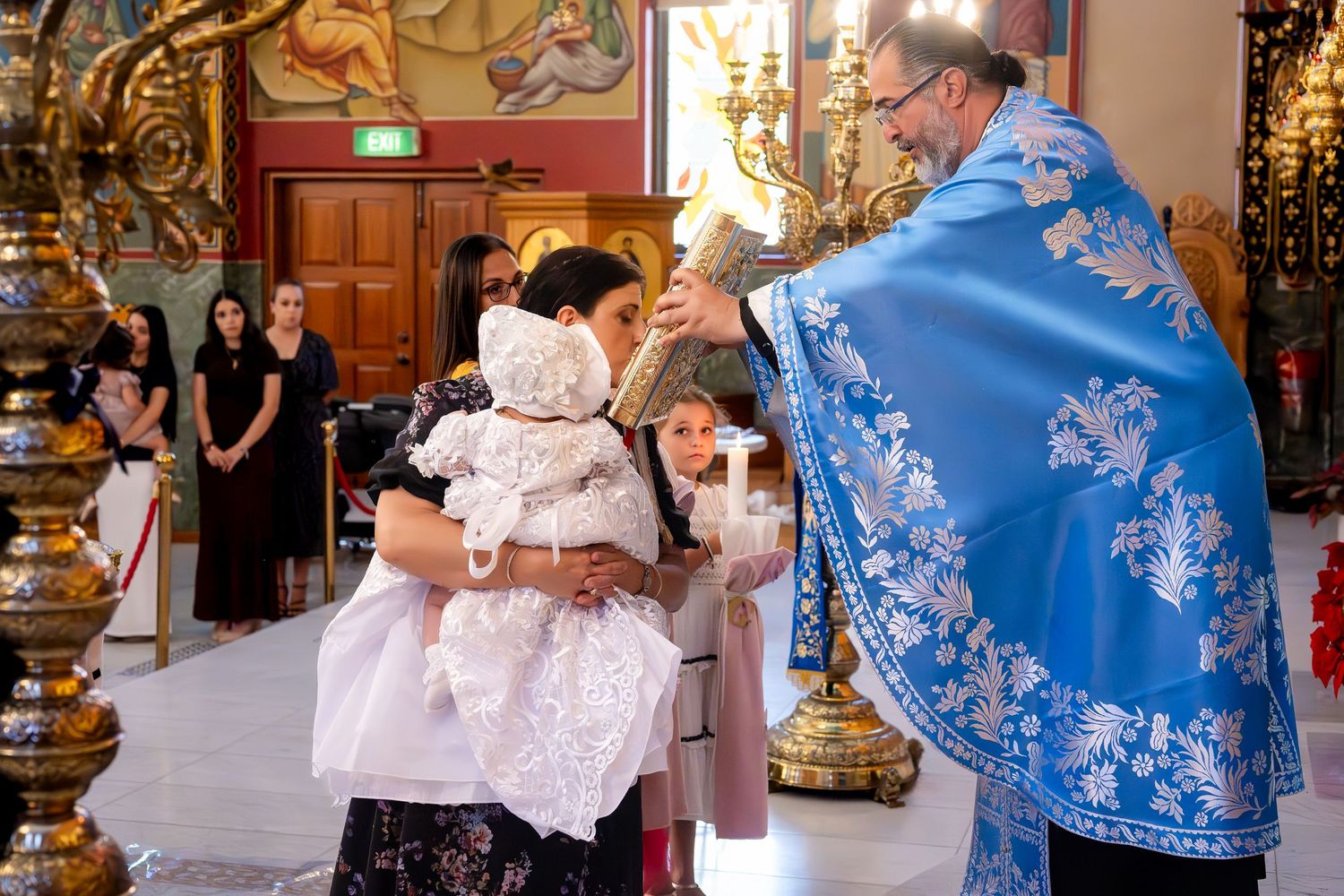 Priest in blue vestments performs a baptism ceremony, blessing an infant dressed in white christening gown inside a church.