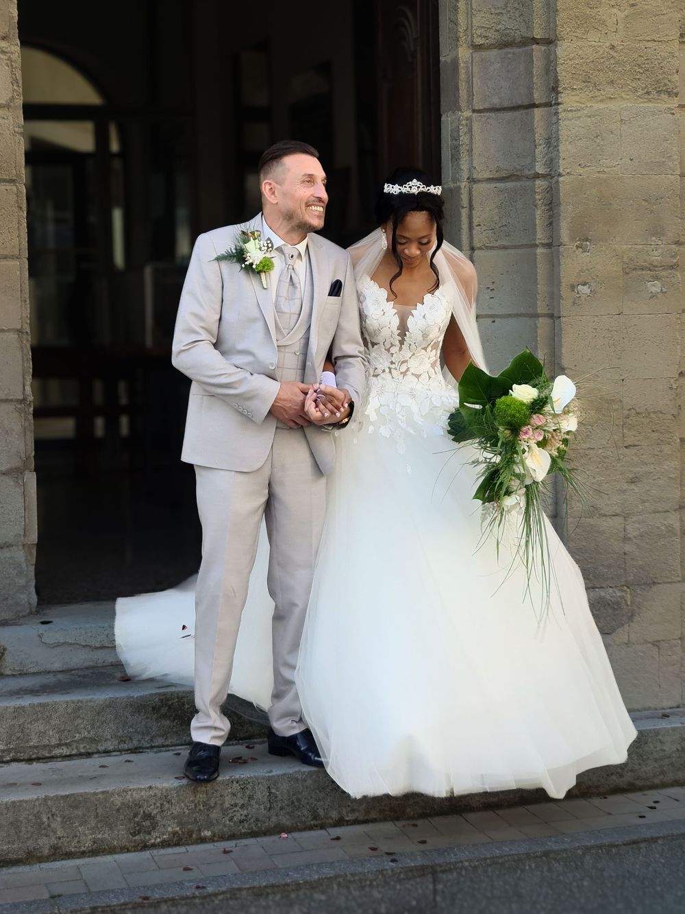Newlywed couple in wedding attire walking down church steps, groom in beige suit, bride in white ballgown holding bouquet.