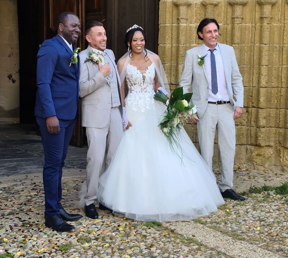 Bride in white mermaid gown holds green bouquet, posing with three suited men outside a church.