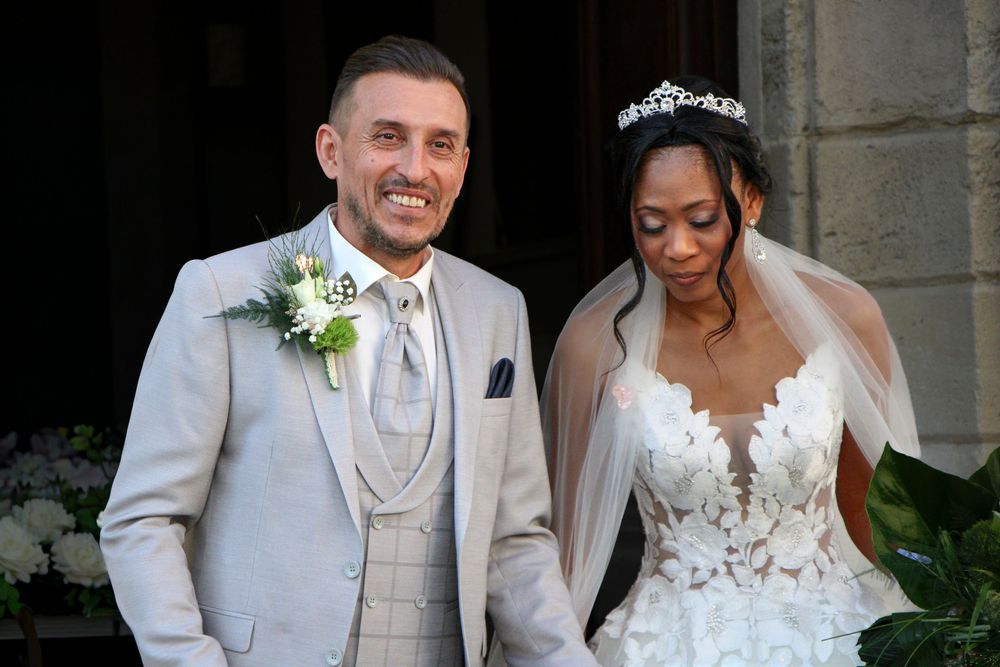 A smiling couple in wedding attire, groom in gray suit and bride in white lace gown with tiara, posing outside a church.
