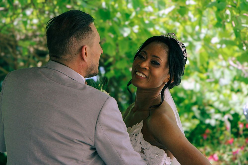 A smiling bride in a white gown gazes lovingly at her groom in a beige suit during an outdoor wedding ceremony.