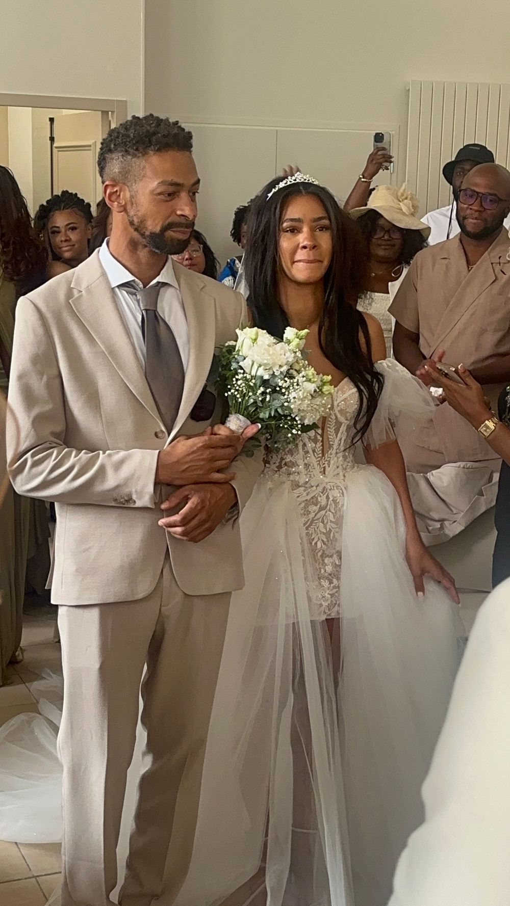 Bride in white lace gown holding bouquet walks down aisle with man in beige suit at wedding ceremony.