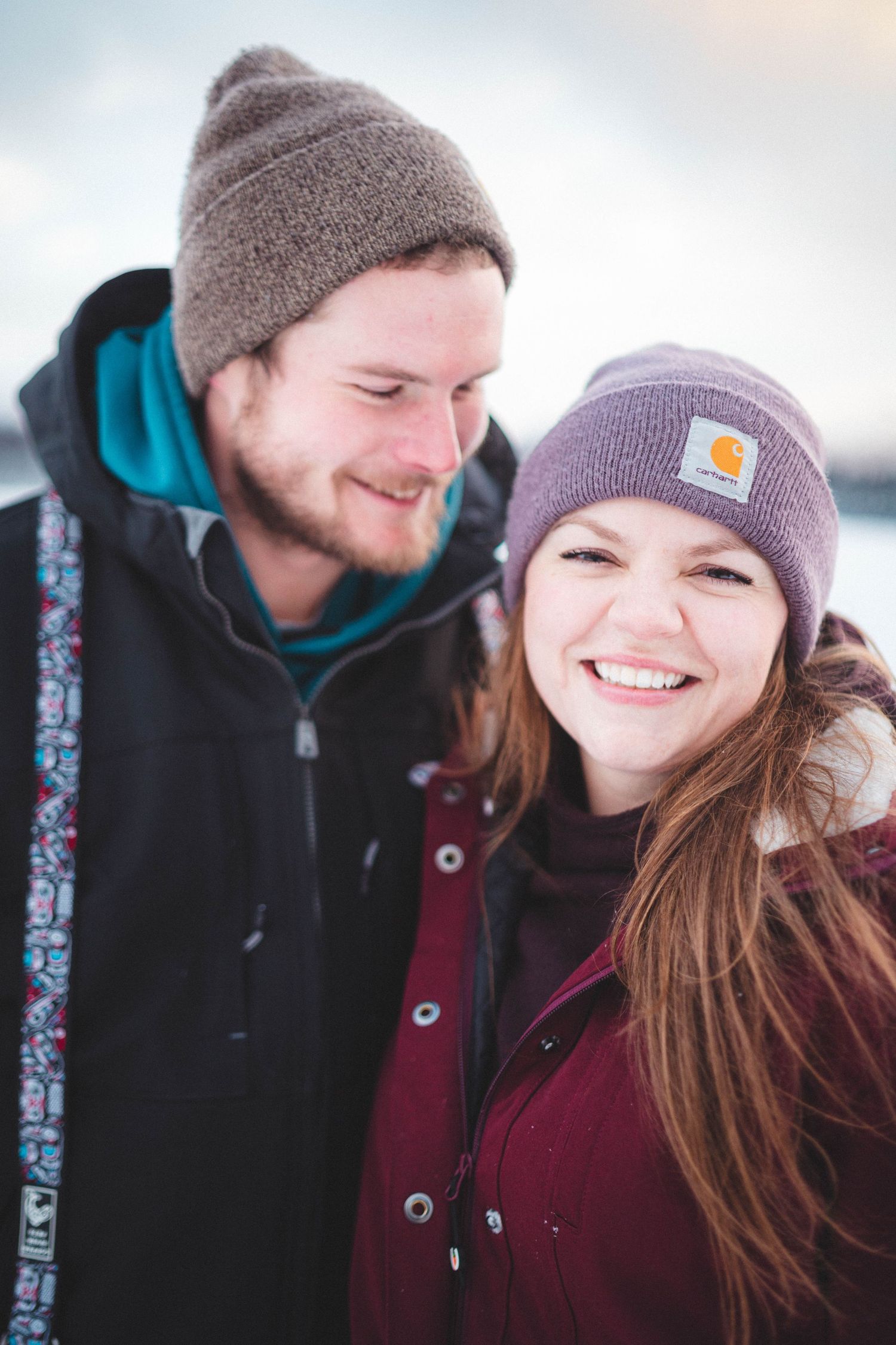 A smiling couple in winter beanies and jackets poses outdoors in a snowy setting, the woman wearing a Carhartt hat.