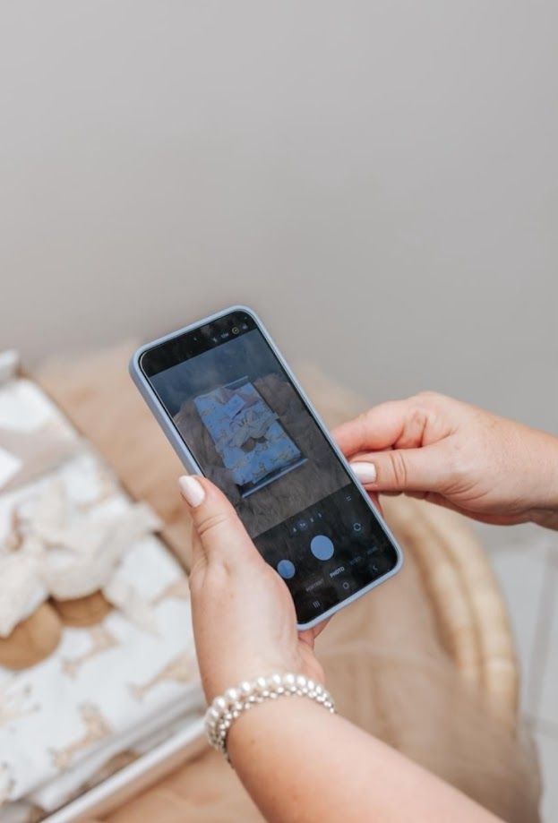 Person with pearl bracelet photographing a blue artwork on a smartphone camera at a white decorative display.