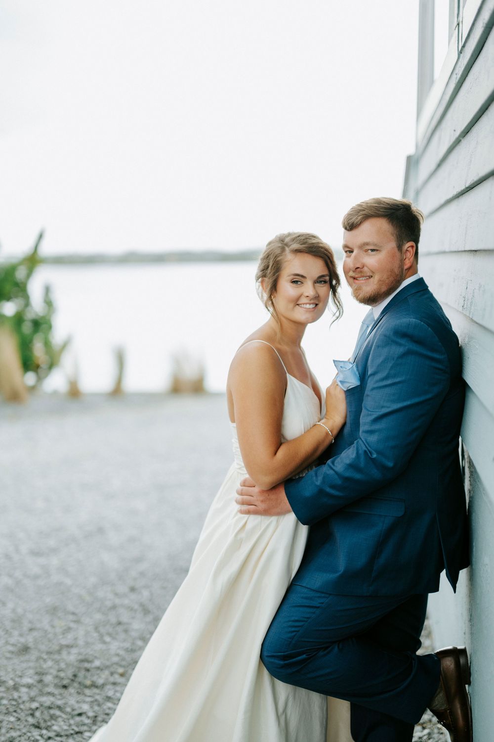 Bride and groom pose together near waterfront building, she in white gown, he in navy suit, smiling warmly.