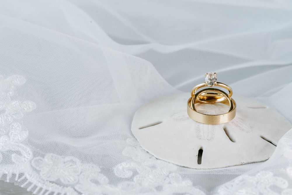 Gold wedding bands and diamond engagement ring displayed on a sand dollar atop a white lace bridal veil.