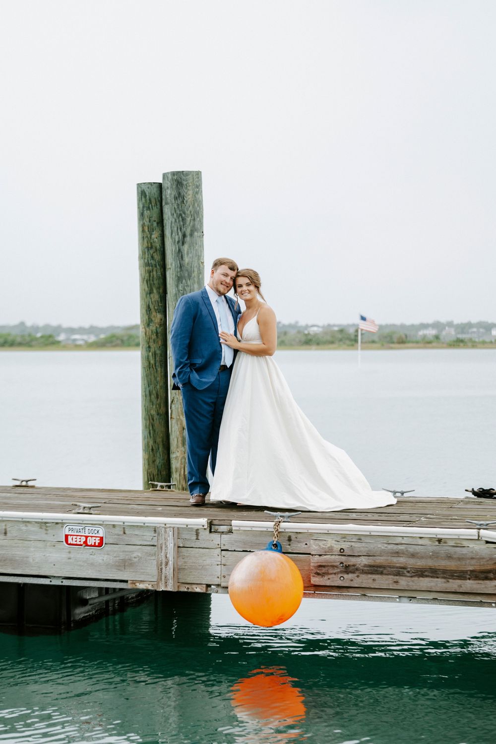 Bride and groom posing on a waterfront dock beside a wooden post, with a white wedding gown and blue suit.