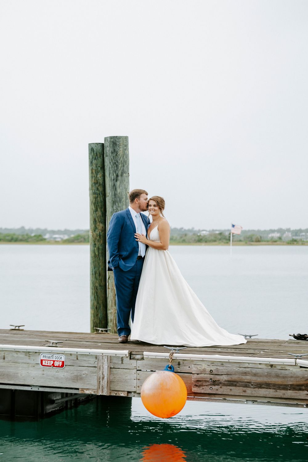 Bride and groom share a romantic moment on a wooden dock by the water, groom in blue suit, bride in flowing white gown.