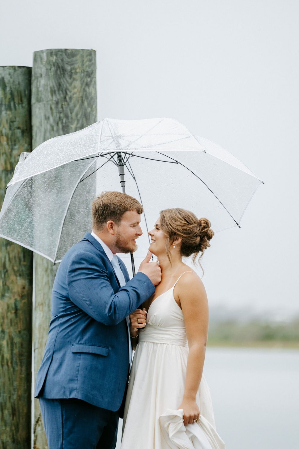 Bride and groom sharing a romantic moment under a clear umbrella on a rainy wedding day by the water.