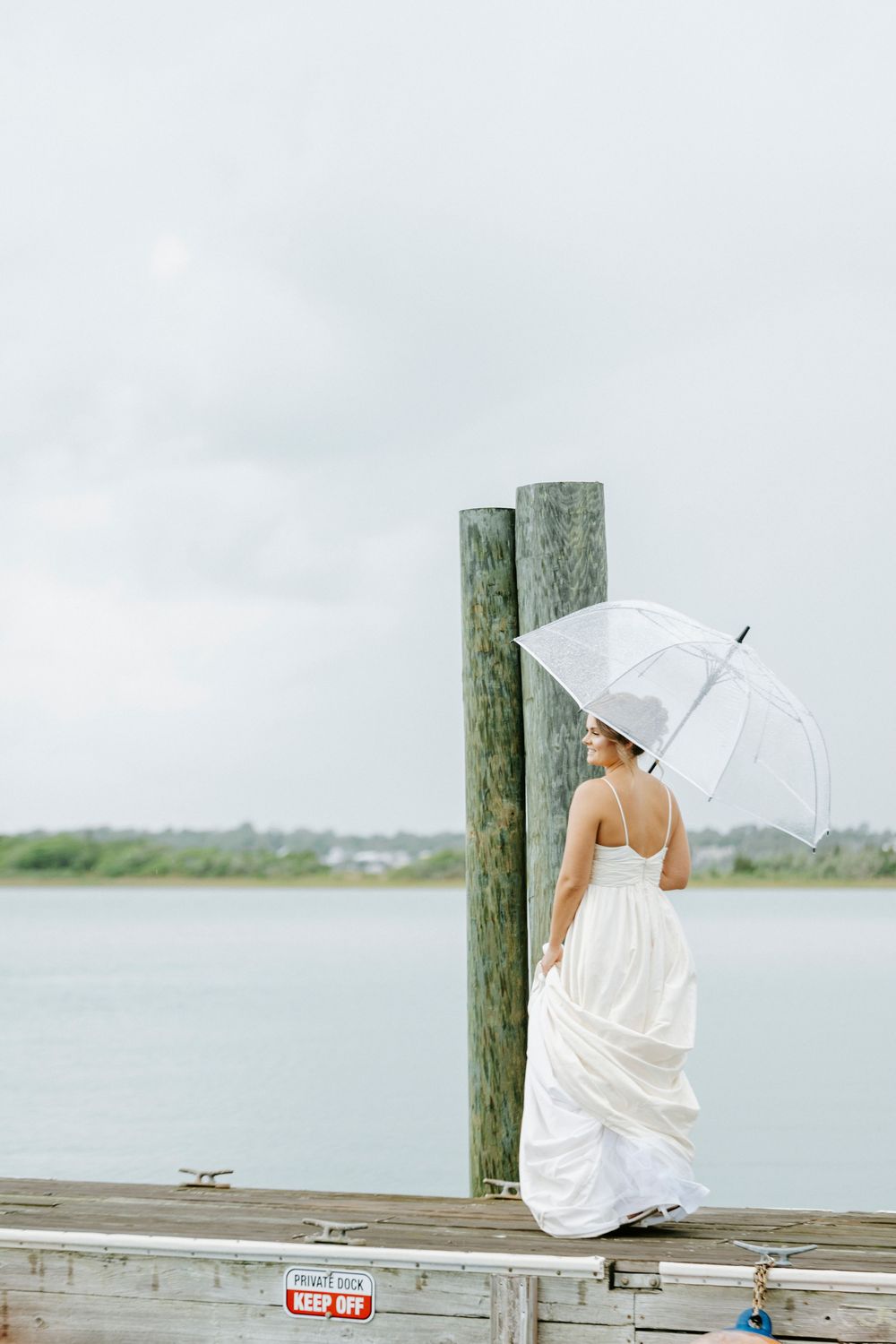 Bride in a white wedding gown holding a clear umbrella while standing on a dock overlooking calm water.